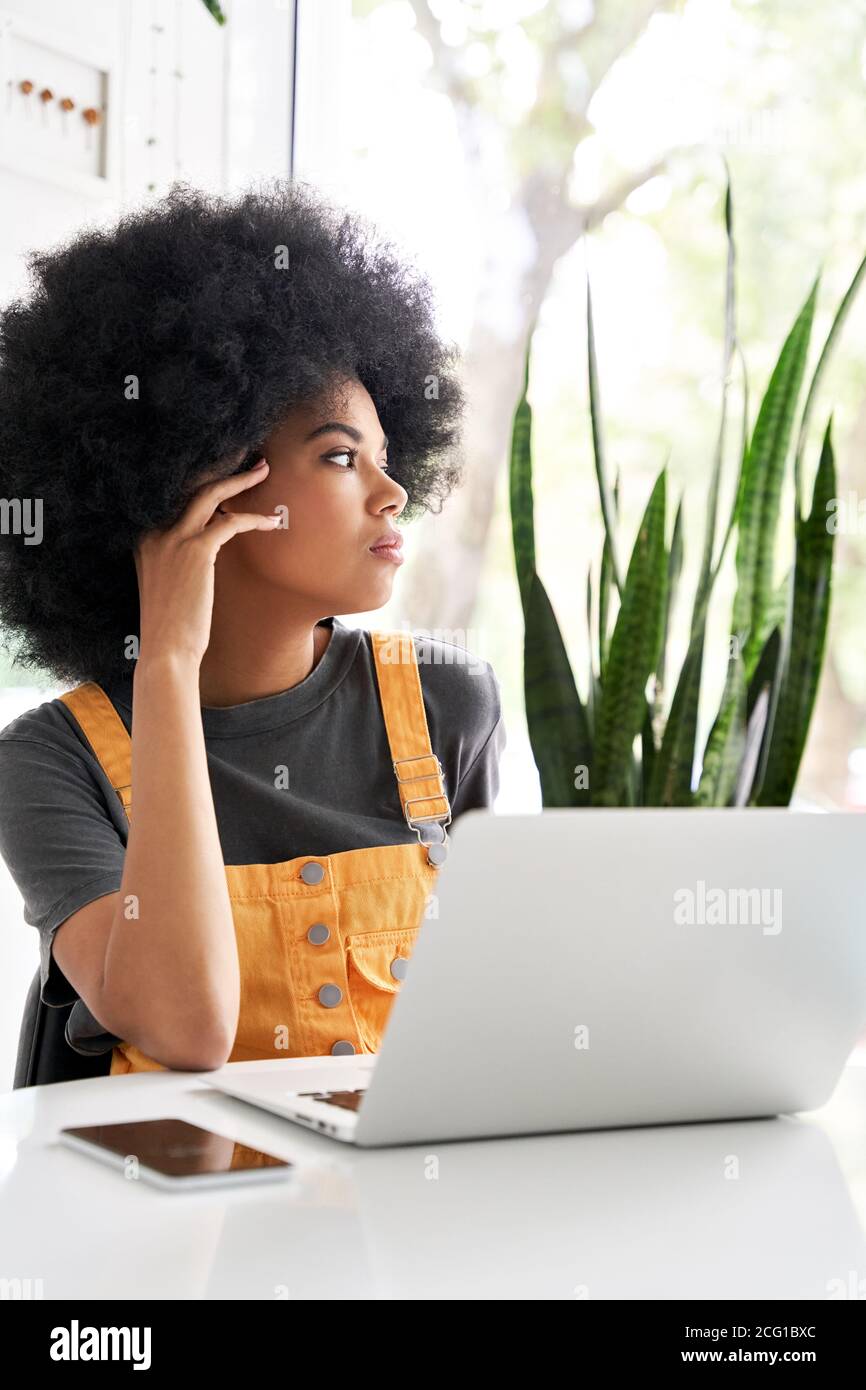 Eine nachdenkliche afrikanische Dame, die durch das Fenster schaute und am Kaffeetisch saß, benutzt einen Laptop. Stockfoto