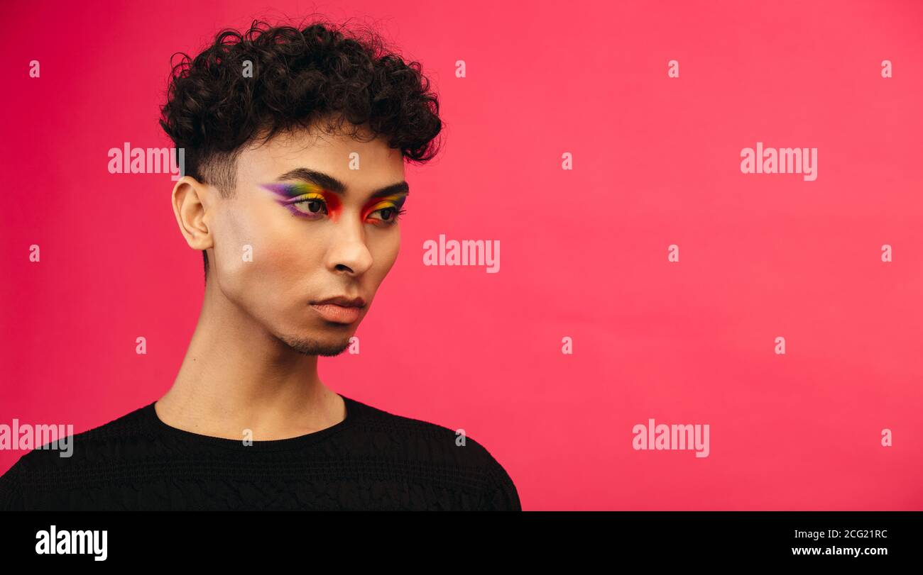 Nahaufnahme eines Geschlechts Flüssigkeit Mann mit Regenbogen Auge Make-up Blick weg auf rotem Hintergrund. Transgender-Mann mit Stolz Flagge Lidschatten. Stockfoto