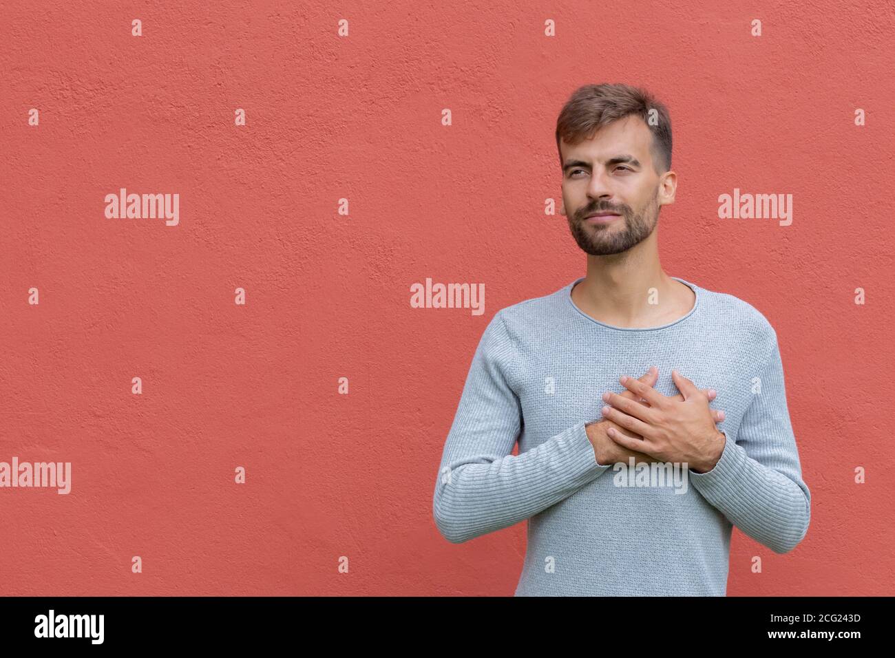 Gutmütiger gutaussehender Kerl hält Hände auf Herz, zeigt Liebe und Sympathie über rote Wand Hintergrund mit Kopieplatz. Körpersprache und Liebesgefühl c Stockfoto