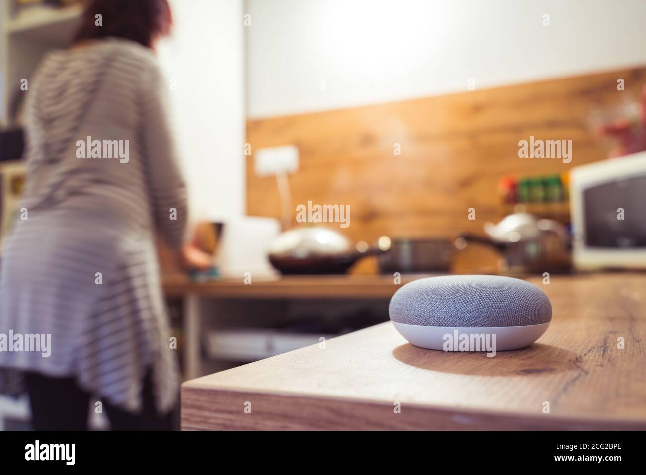 Sprachgesteuerter Smart Speaker in einem Zuhause mit Sprachsteuerung Smart Speaker in einer Innenraumumgebung mit einer Frau Kochen im Hintergrund Stockfoto