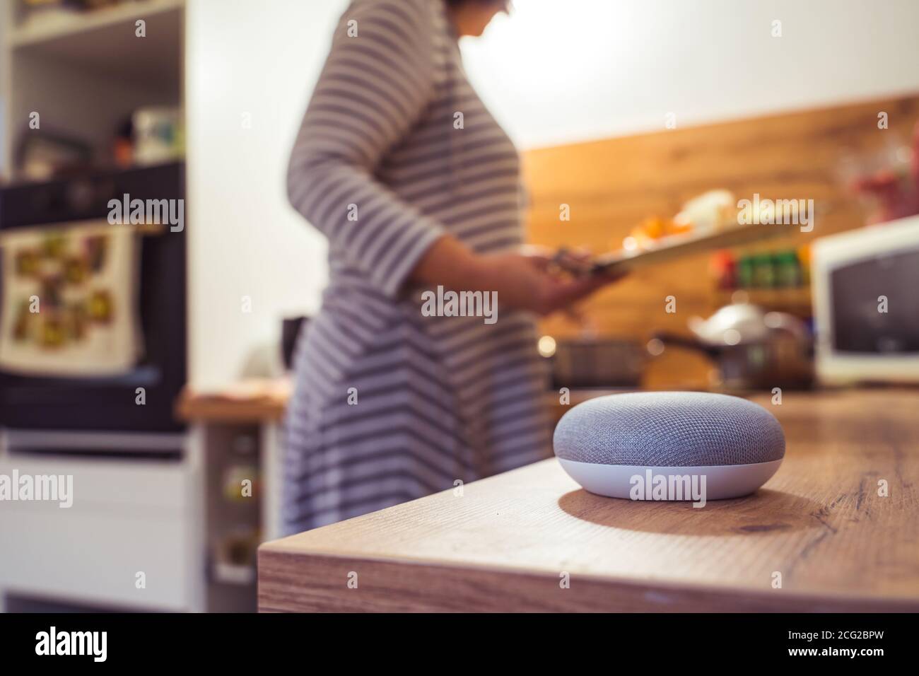 Sprachgesteuerter Smart Speaker in einem Zuhause mit Sprachsteuerung Smart Speaker in einer Innenraumumgebung mit einer Frau Kochen im Hintergrund Stockfoto