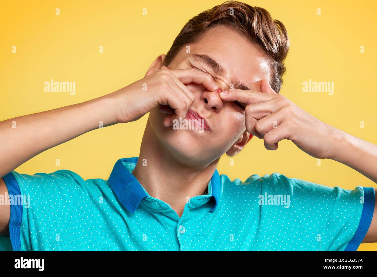 Kosmetologie, Dermatologie und Akne. Ein Teenager in einem blauen T-Shirt quetscht mit den Fingern einen Pickel auf seine Nase, die Augen sind vor Schmerzen geschlossen. Gelbe Rückenfarbe Stockfoto