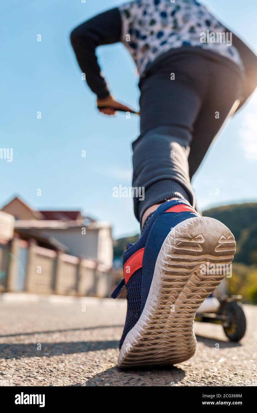 Ein Teenager fährt einen Roller. Linker Fuß in Nahaufnahme eines Sneakers. Ansicht von unten und hinten. Leere Straße bei sonnigem Wetter im Hintergrund. Konzept des Outs Stockfoto
