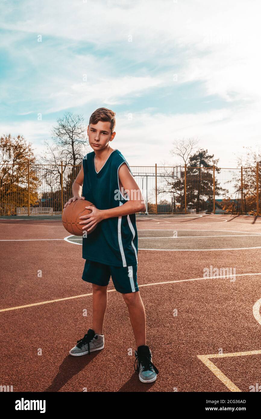 Sport und Basketball. Ein junger Teenager in einem blauen Trainingsanzug  posiert mit einem Basketball in den Händen. Im Hintergrund ein  Basketballplatz. Vertikal. Cop Stockfotografie - Alamy