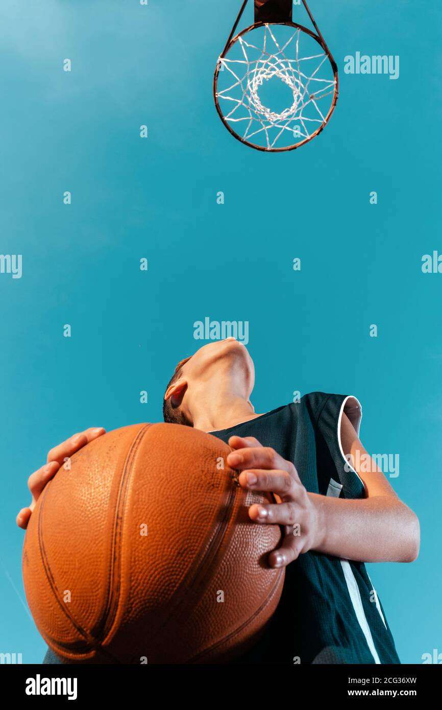 Sport und Basketball. Ein junger Teenager im schwarzen Trainingsanzug steht mit einem Ball in den Händen und bereitet sich darauf vor, den Ball in den Ring zu werfen. Blauer Himmel wi Stockfoto