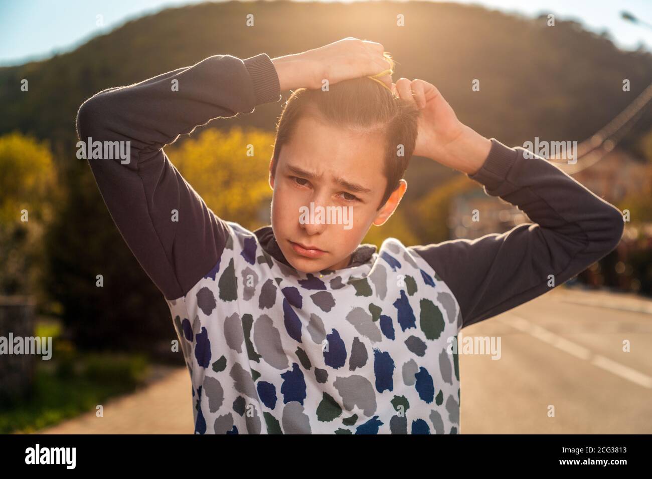 Ein Teenager bindet seine Haare mit einem peinlichen Blick in einen Pferdeschwanz. Emotionen. Straße im Hintergrund. Das Konzept der Frisuren und Selbstpflege. Stockfoto