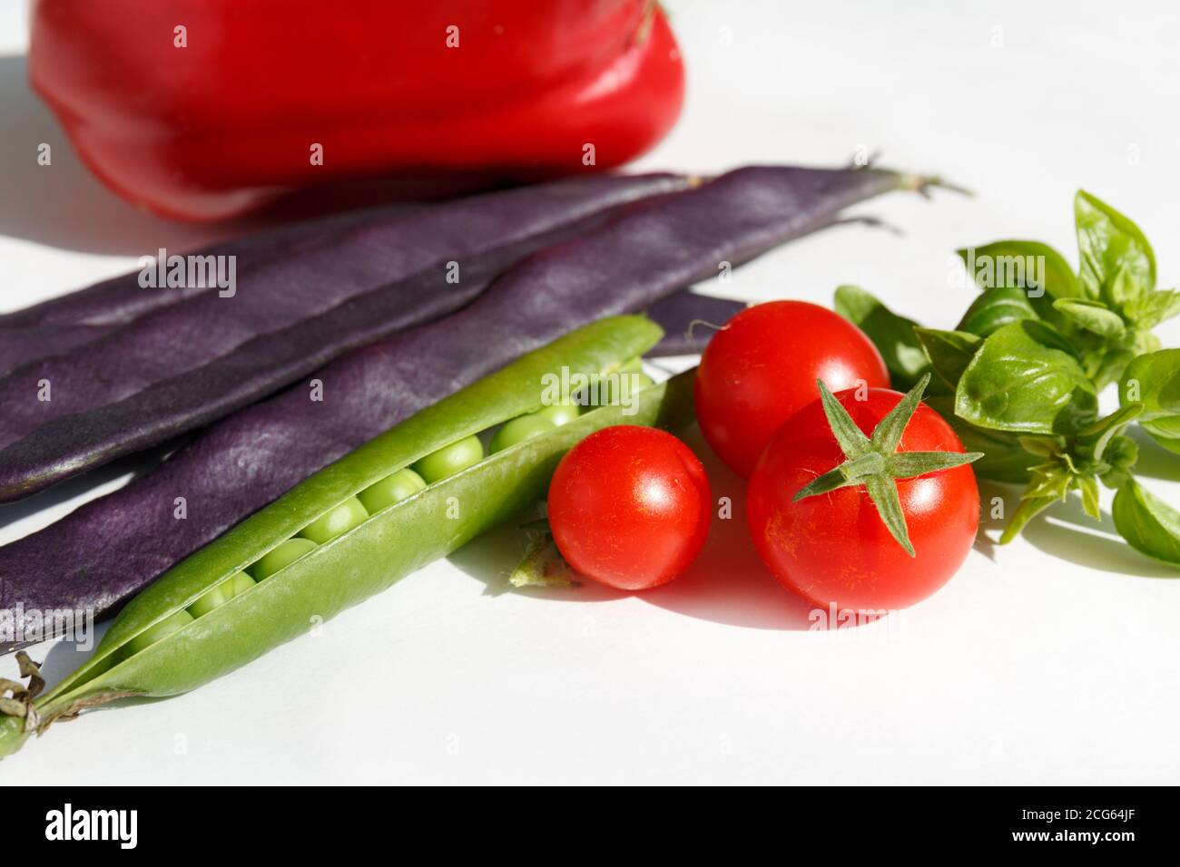 Kirschtomaten, grüne Basilikumblätter, eine offene Erbsenschote, violette Bohnenschoten und roter Paprika. Saisonales Gemüse auf weißem Hintergrund. Herbsternte Stockfoto
