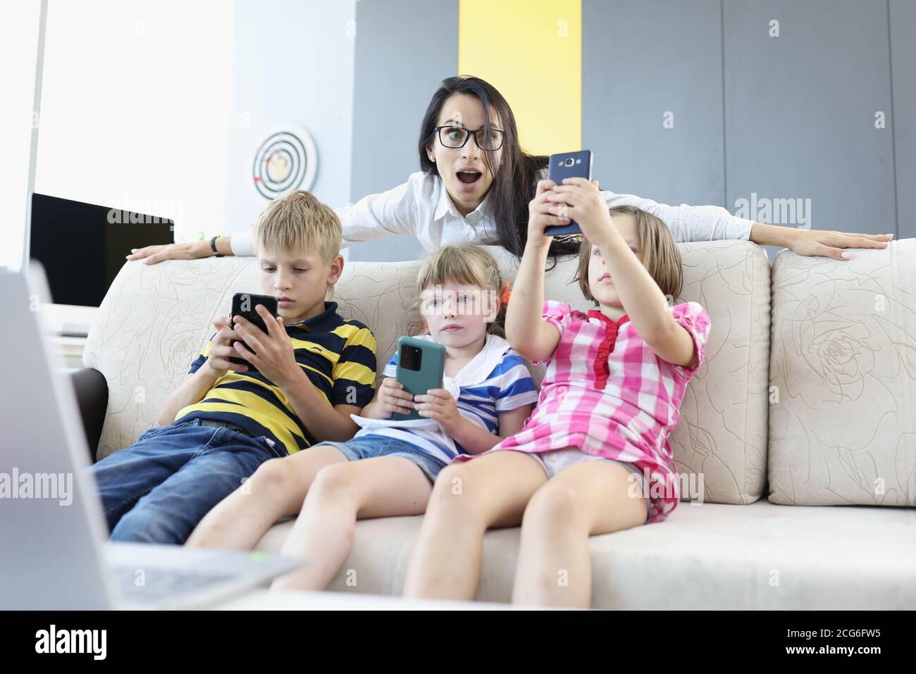 Drei Kinder sitzen auf der Couch mit Smartphones in den Händen, spielen Online-Spiele mit Frau hinter der Couch stehen und Blick erschrocken Stockfoto