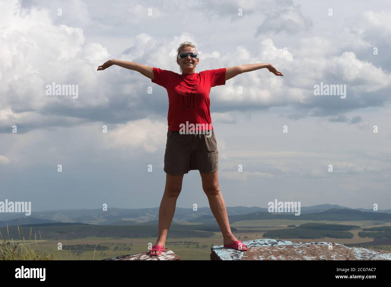 Eine Erwachsene Frau steht mit ausgestreckten Armen auf einem hohen Berg vor dem Hintergrund einer Ebene und eines wolkigen Himmels. Stockfoto