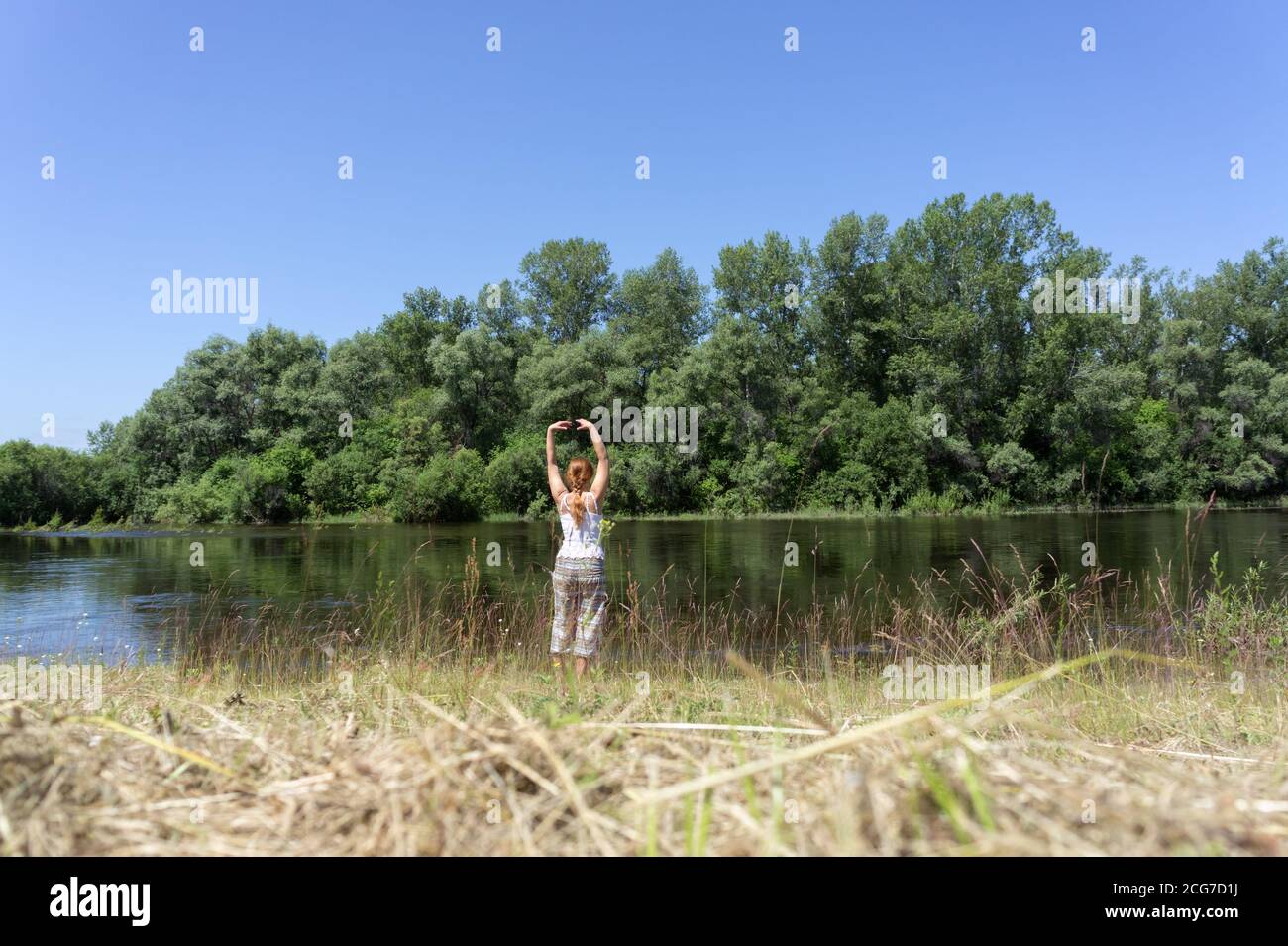 Das rothaarige Mädchen, das in der Yoga-Gymnastik am Ufer des Flusses auf dem Hintergrund der Bäume an einem sonnigen Sommertag beschäftigt ist. Stockfoto