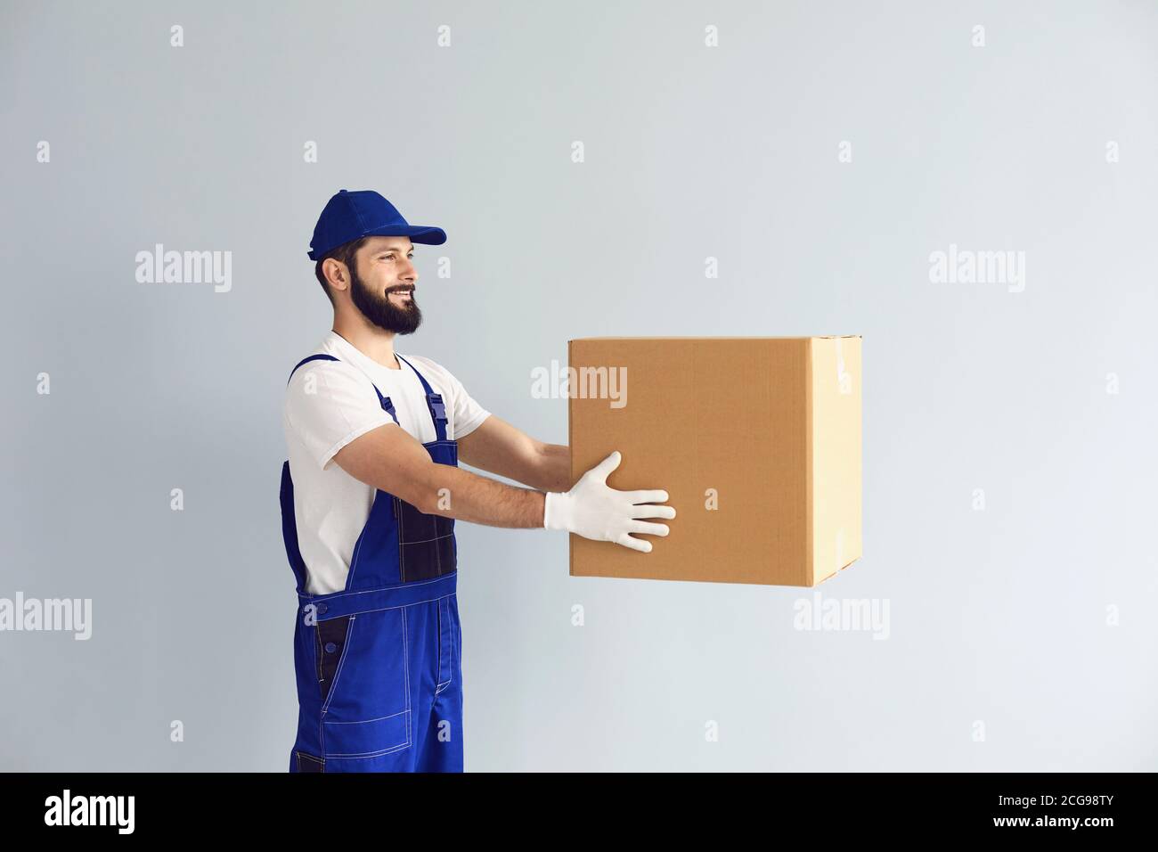 Courier hält die Kiste vor sich hin und übergibt sie mit einem Lächeln auf grauem Hintergrund. Stockfoto