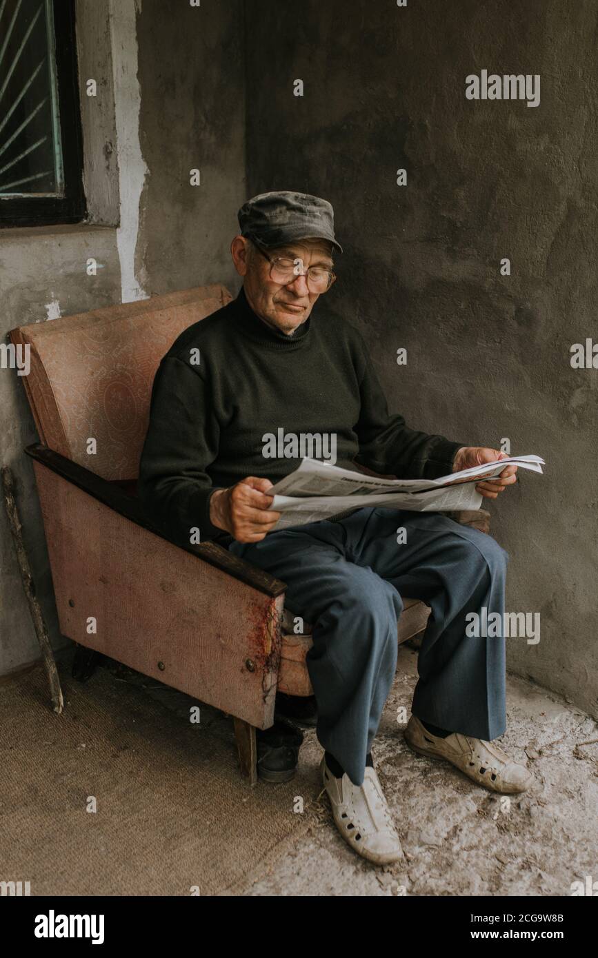 Ein nachdenklicher alter Mann in Gläsern mit grauen Haaren sitzt auf einem alten Sessel und liest eine Zeitung in der Nähe des Hauses. Falten. Weisheit. Gegen eine dunkle GRA Stockfoto