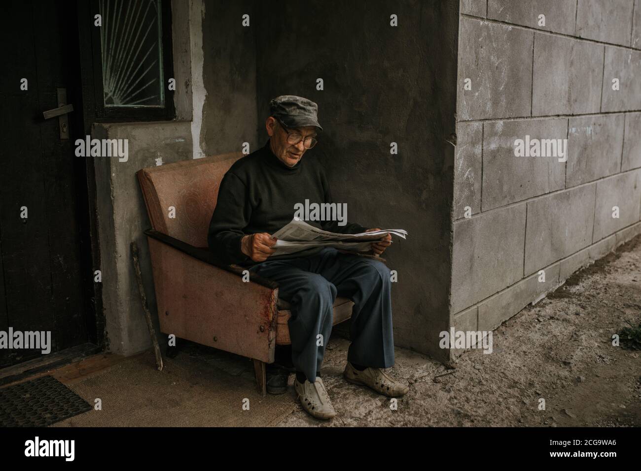Ein nachdenklicher alter Mann in Gläsern mit grauen Haaren sitzt auf einem alten Sessel und liest eine Zeitung in der Nähe des Hauses. Falten. Weisheit. Gegen eine dunkle GRA Stockfoto