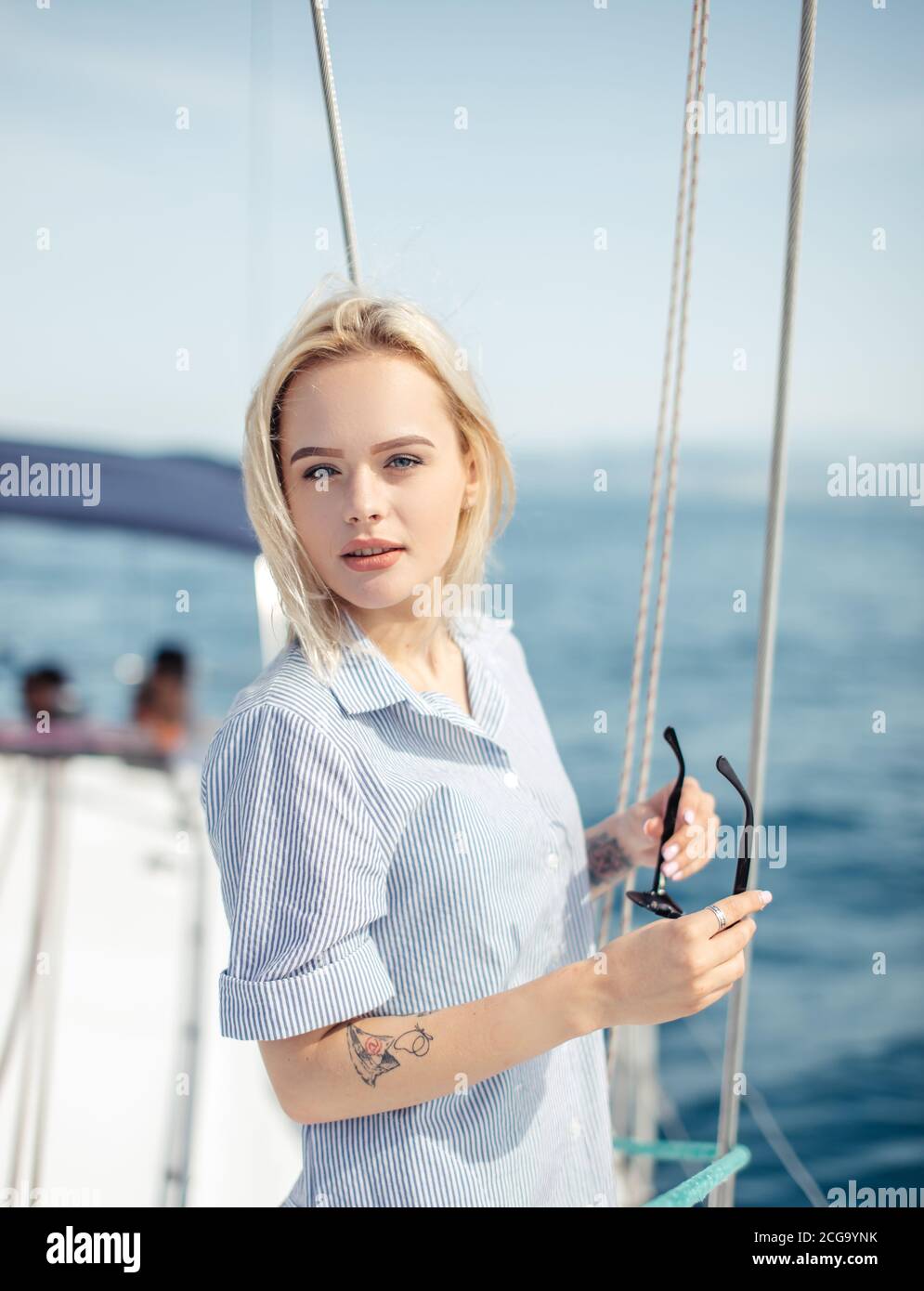 Außenportrait der schönen jungen Frau, die auf dem Deck eines Segelbootes mit erhöhten Segeln steht und auf die Kamera schaut Stockfoto