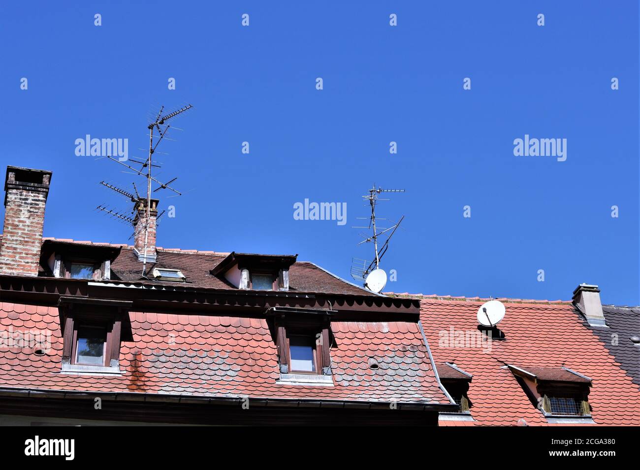 Rot-braun gefliestes Verkleidungen-Dach mit Dachfenstern oder Dachfenstern, Parabeln und altmodischen analogen TV-Antennen in Colmar, Frankreich unter blauem Himmel. Stockfoto