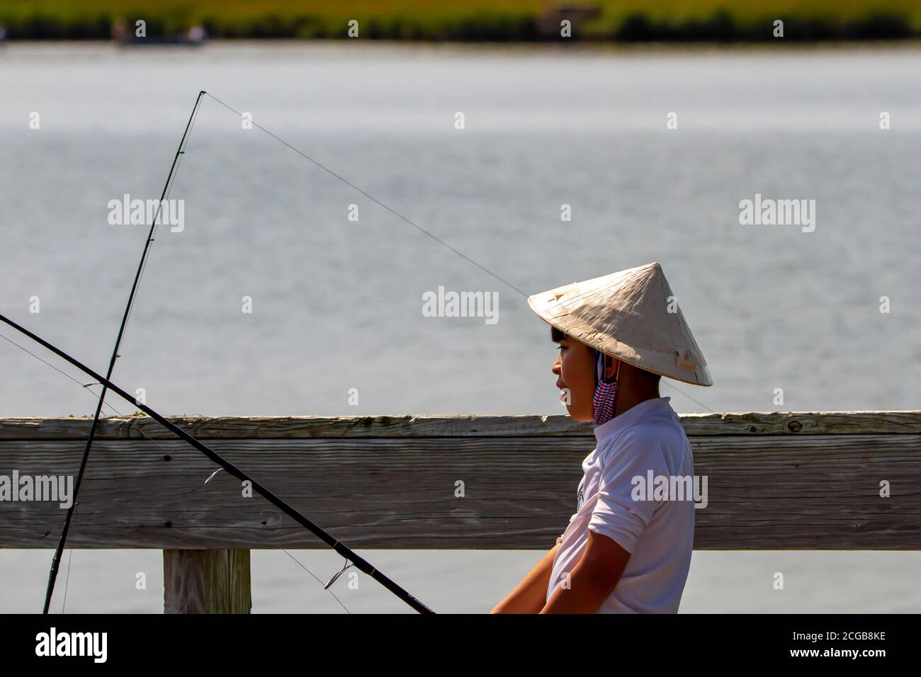 Eastern Neck Island, MD, USA 09/06/2020: Ein asiatischer Junge mit einem traditionellen chinesischen Strohbambus-Coolie-Kegelhut hält eine Angelrute auf einer Brücke Stockfoto