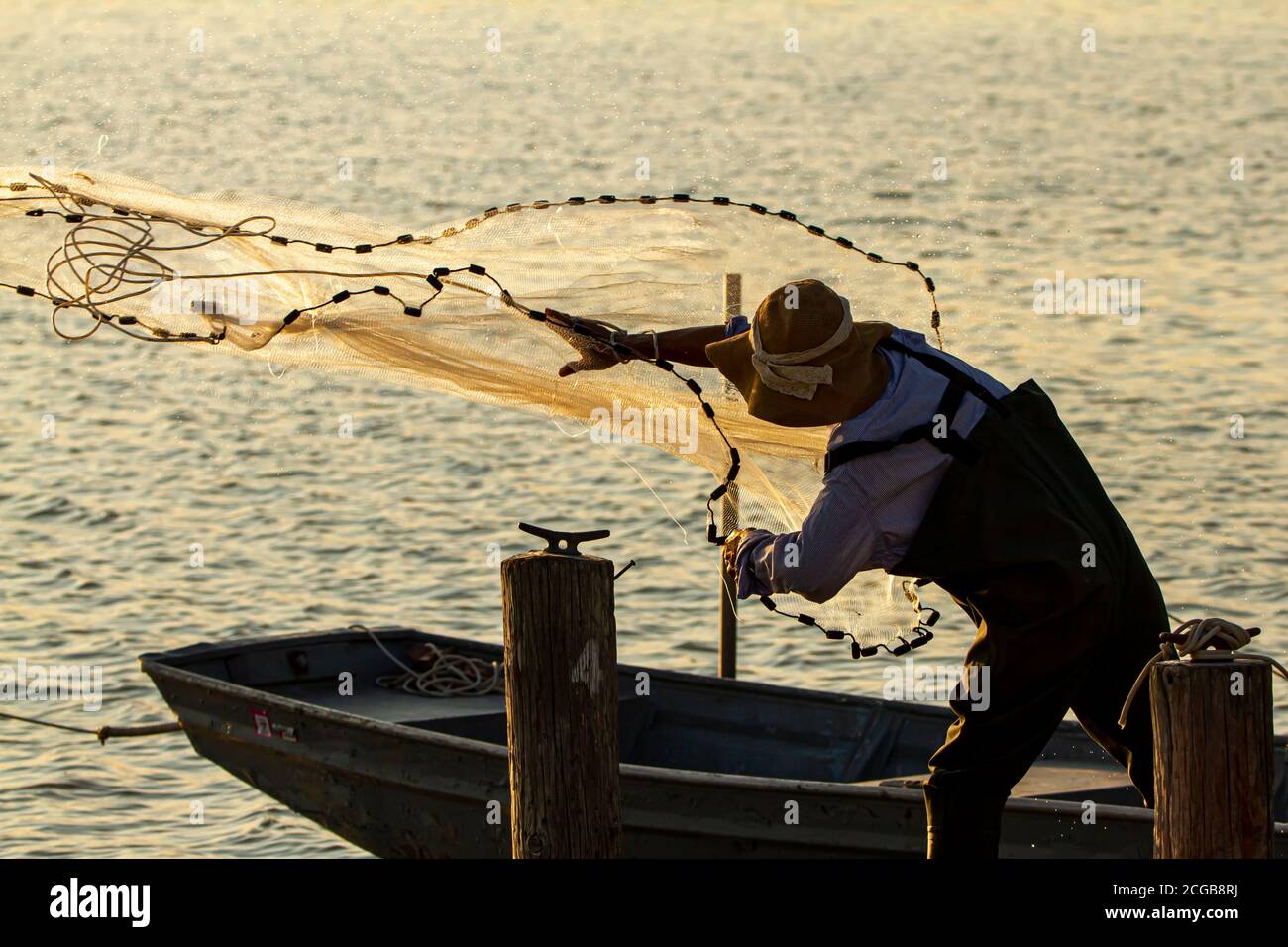 Ein Fischer mit Overalls und Stiefeln sowie einem Eimerhut wirft bei Sonnenuntergang ein Wurfnetz von einem hölzernen Pier ins Meer. Er ist hinterleuchtet und sieht Stockfoto