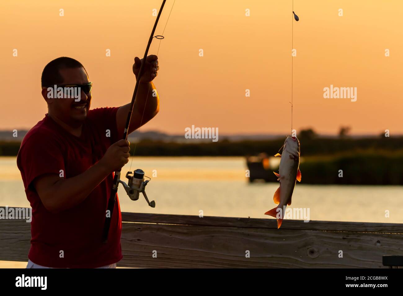 Eastern Neck Island, MD, USA 09/06/2020: Ein junger hispanic-Mann mit rotem T-Shirt und Sonnenbrille hat gerade einen Katzenfisch mit seiner Angelrute gefangen. Er Stockfoto