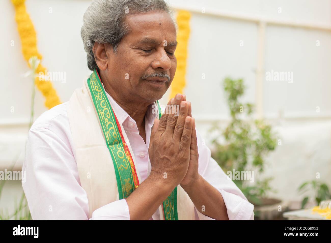 Älterer Mann in geschlossenen Augen bietet Gebet durch Namaste Geste beten zu gott zu Hause in traditioneller Kleidung während Ritualen. Stockfoto