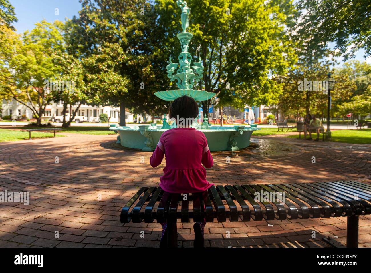 Ein kleines Mädchen in pinker Freizeitkleidung sitzt allein auf einer Metallbank im Fountain Park von Chestertown, Maryland. Im unscharfen Hintergrund Stockfoto