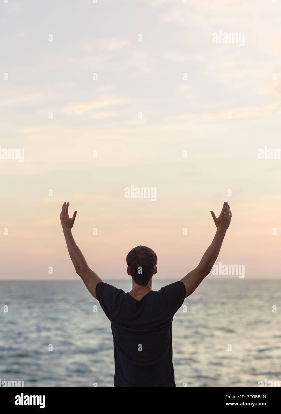 Gesunder junger Mann mit den Armen nach oben zum Himmel, der während eines wunderschönen Sonnenuntergangs mit Blick auf den Ozean feiert. Wohlbefinden und Freiheit. Stockfoto