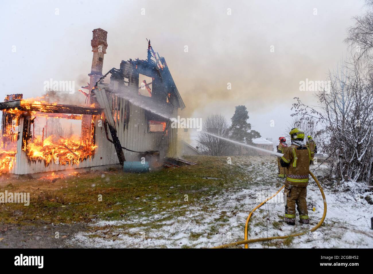 Norwegischer Feuerwehrmann von hinten versucht, Flammen auf Haus in Brand mit riesigen Flammen und Rauch, im Winter mit Schnee zu löschen. Stockfoto