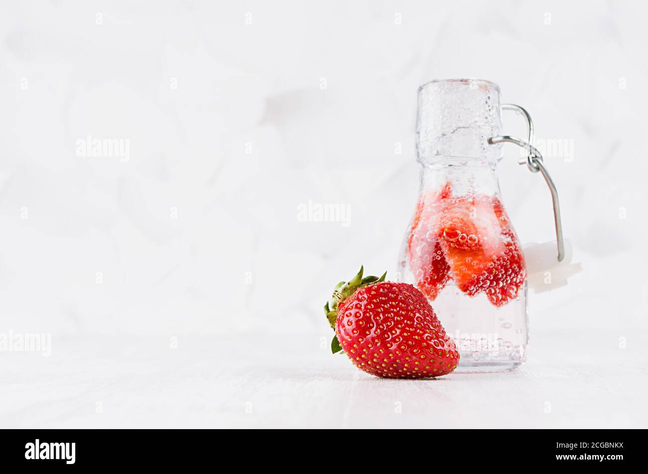 Erfrischendes kaltes Fruchtgetränk mit roter Erdbeere, helle Blasen in transparenter Yoke-Flasche im eleganten weißen Interieur auf Holztisch, Kopierraum. Stockfoto