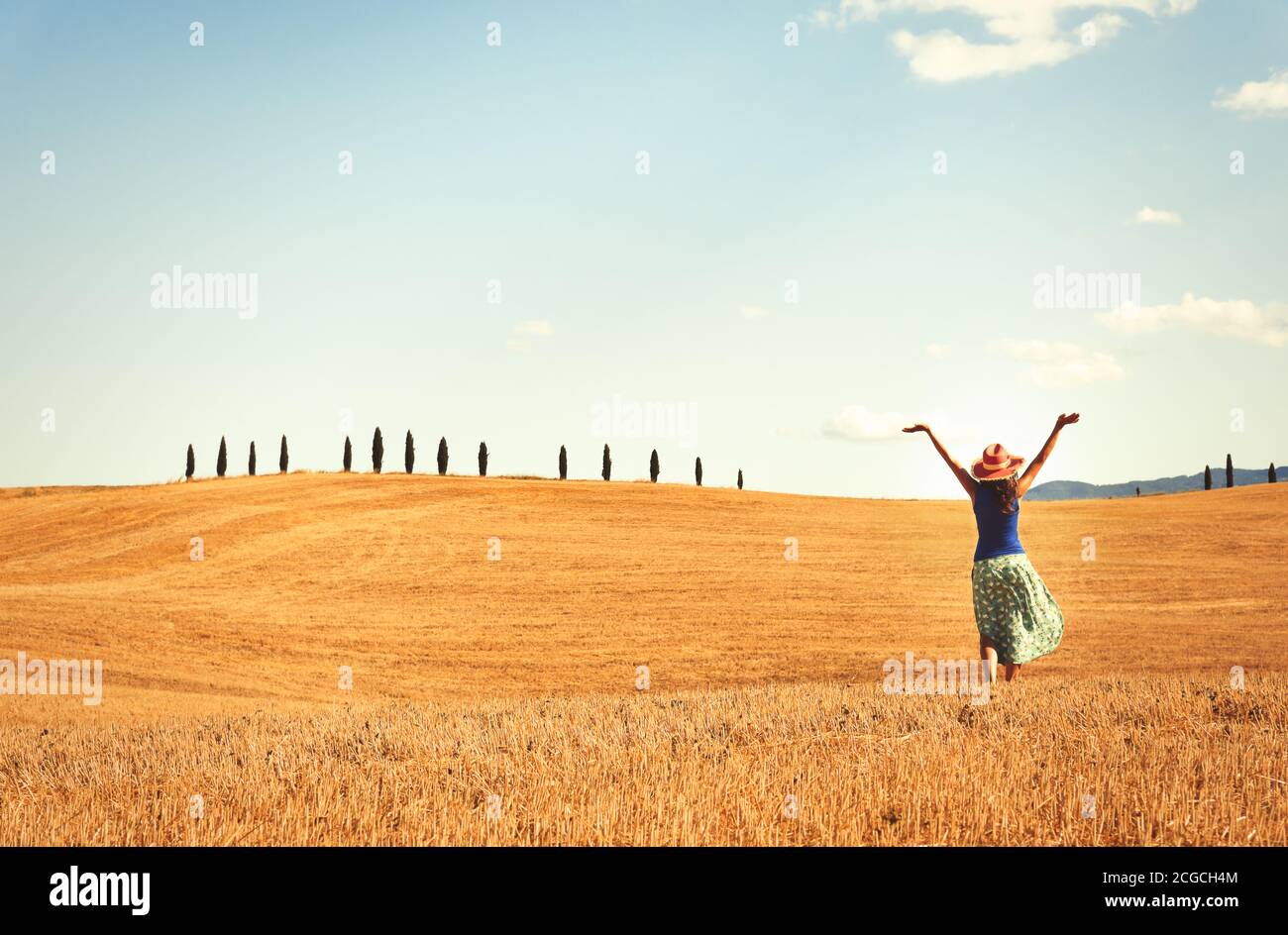 Glückliche Frau genießen die Freiheit, draußen in einem Weizenfeld. Zypressen am Horizont mit blauem Himmel und weißem Wolkenhintergrund. Stockfoto