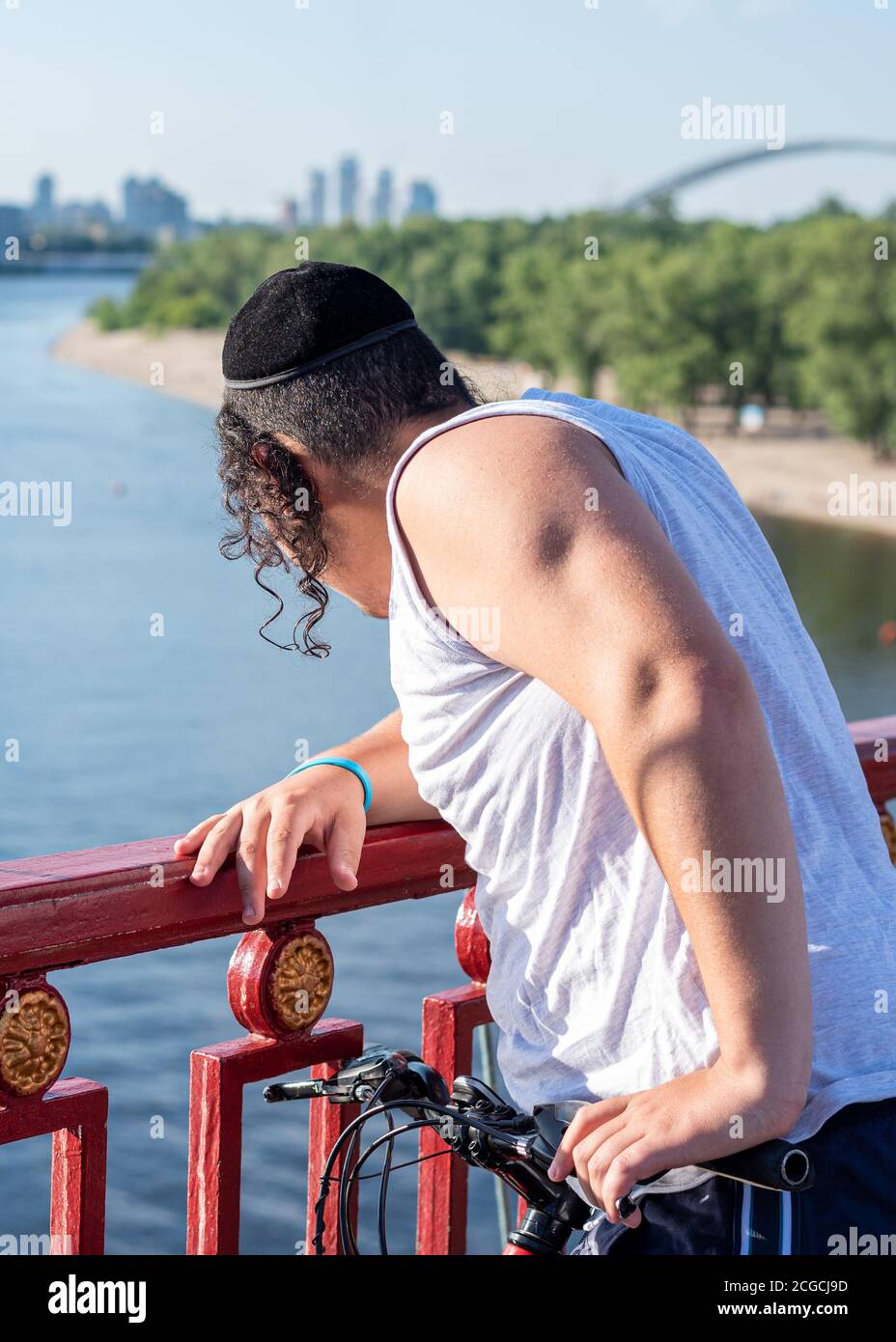 Nicht erkennbarer junger jüdischer Mann mit verdrehtem payot in schwarzem Samtkippah, der auf der Brücke steht und in den Fluss hinunterblickt. Vertikale Ausrichtung Stockfoto