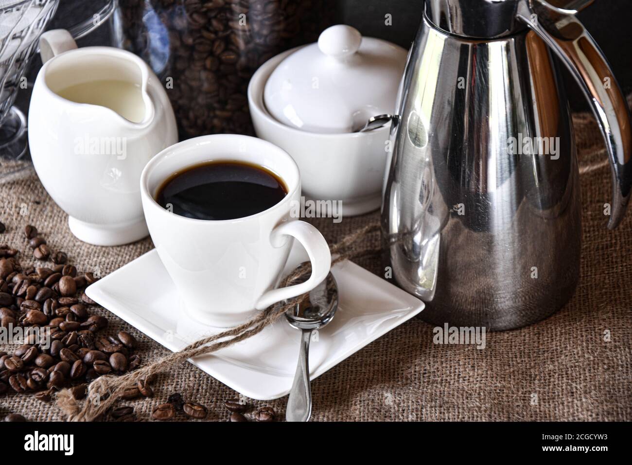 Eine Tasse Kaffee mit weißen Gerichten, ein Milchkännchen und eine Zuckerkanne auf einer Jute-Textur mit silbernen Thermoskanne serviert Kaffeekanne Stockfoto