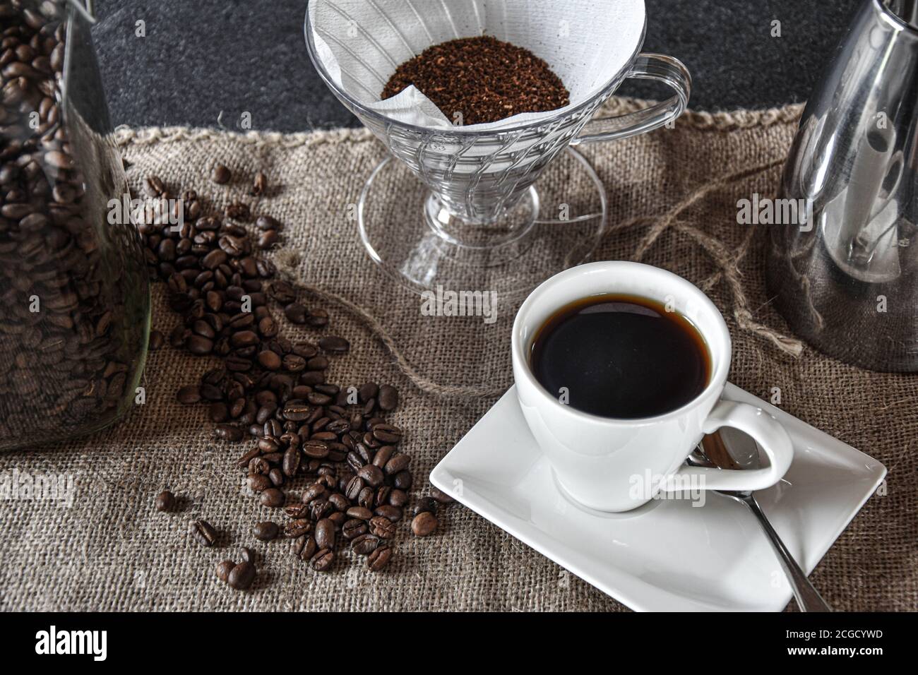 Kaffee in einer weißen Tasse auf einem Jutetuch mit silberner Kaffeekanne, Filter und Kaffeebohnen zur Dekoration Stockfoto