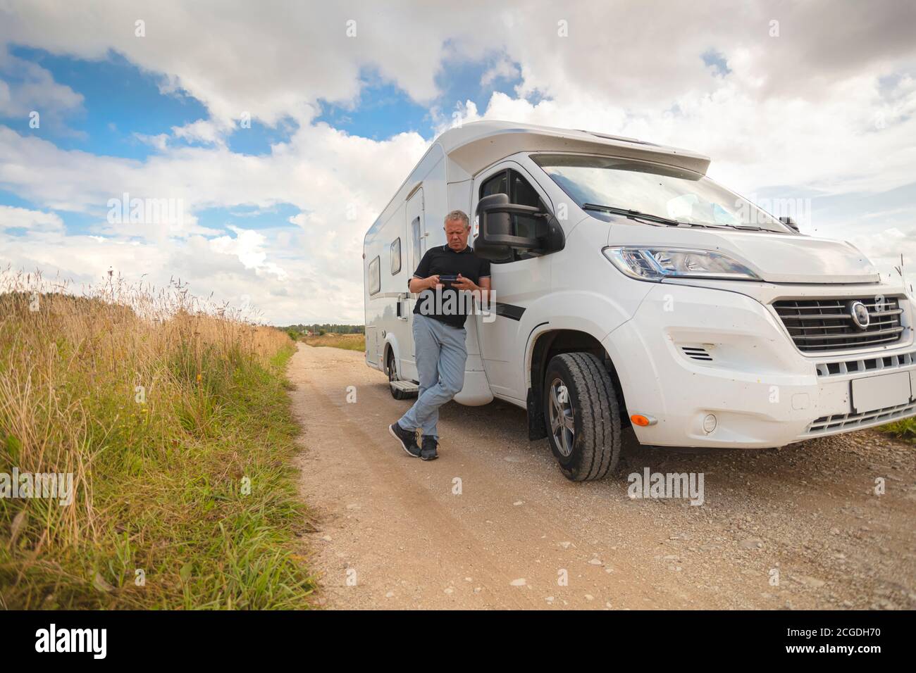 Ein Mann schaut auf das Telefon an einem Wohnmobil auf einer Landstraße mitten auf Feldern. Sommer sonnigen Tag. Hochwertige Fotos Stockfoto