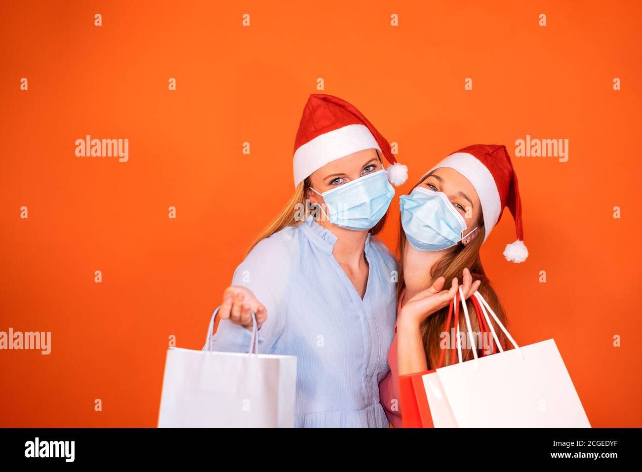 Portrait von zwei Mädchen, die auf die Kamera mit einem Blick Weihnachtshut und medizinische Maske mit farbigen Einkaufstaschen Stockfoto