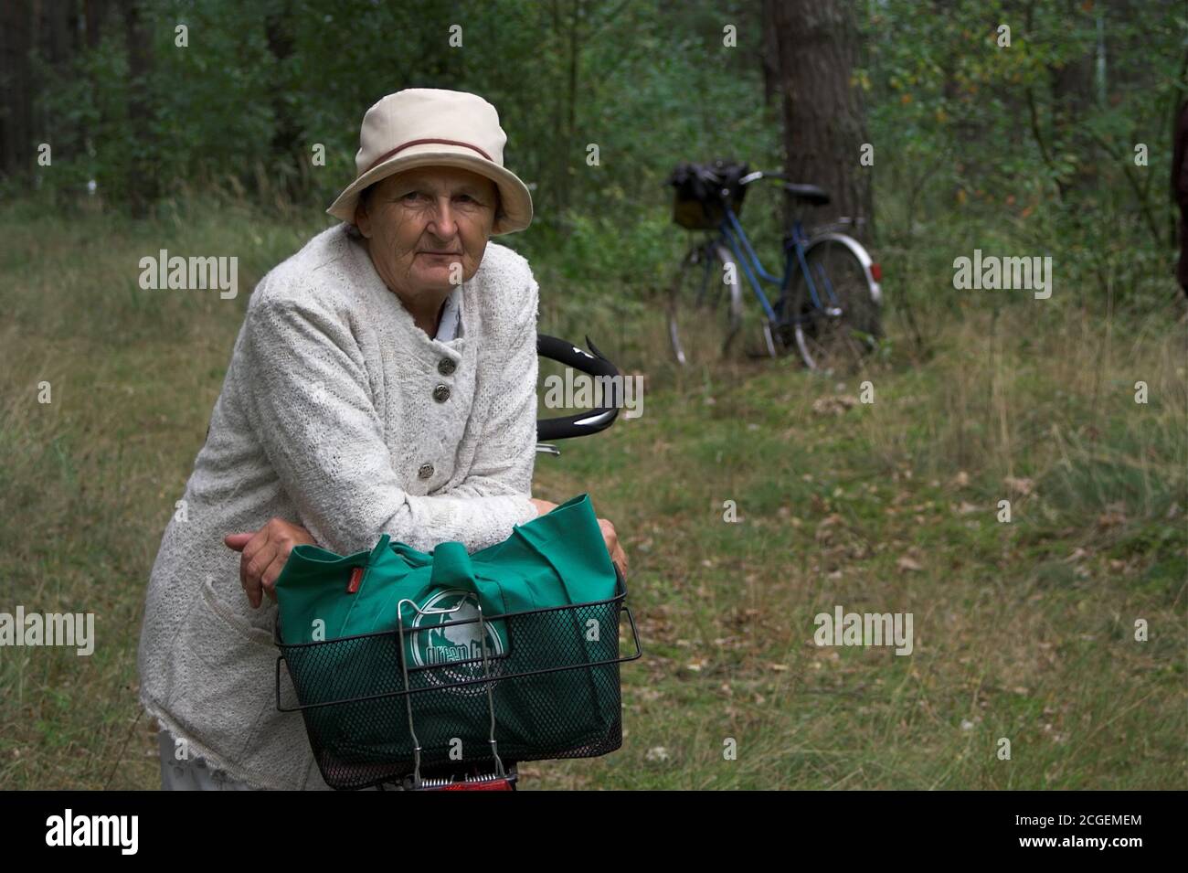 Eine alte Dame im Hut auf einer Wanderung im Wald, lehnt sich an ihr Fahrrad. Eine alte Dame mit Hut, die im Wald wandert und sich an ihr Fahrrad führt. Stockfoto