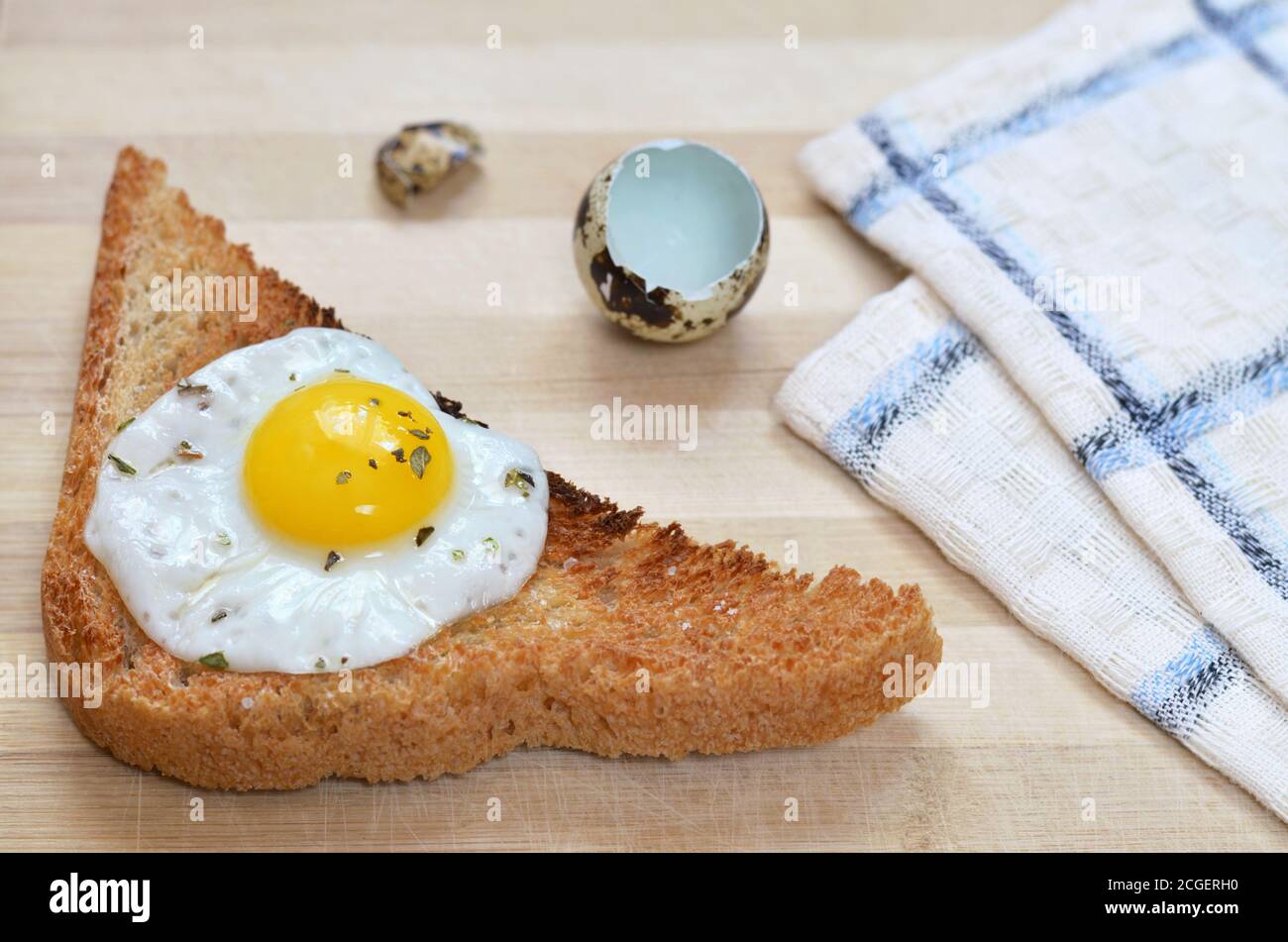 Geröstetes Brot mit gebratenem Wachtelei auf einem hölzernen Schneidebrett mit Küchentuch und Eierschalen, selektiver Fokus. Gesundes Frühstück. Stockfoto