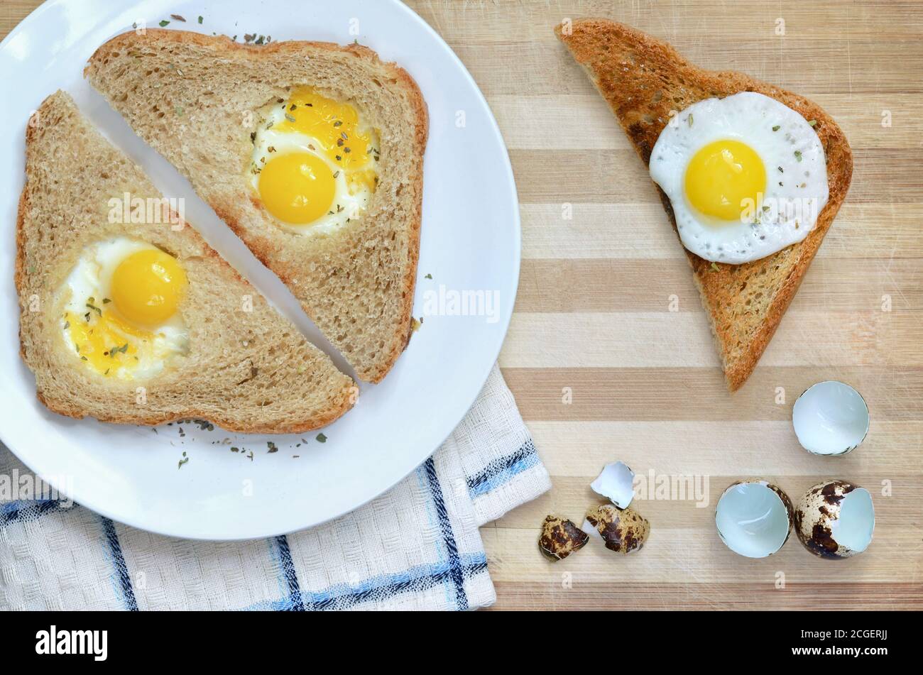 Gebratene Brotscheiben mit Wachteleiern auf einem weißen Teller auf einem hölzernen Schneidebrett mit Küchentuch und Eierschalen, Draufsicht. Stockfoto