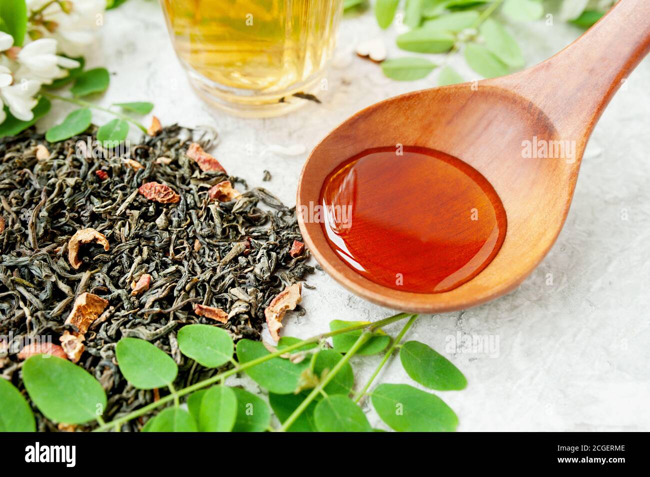 Holzlöffel mit Honig, getrocknete Teeblätter mit Beeren und Akazienblüten Brühe in Glas Tasse mit üppig blühenden weißen Akazienbaum Zweig, nützlich Stockfoto