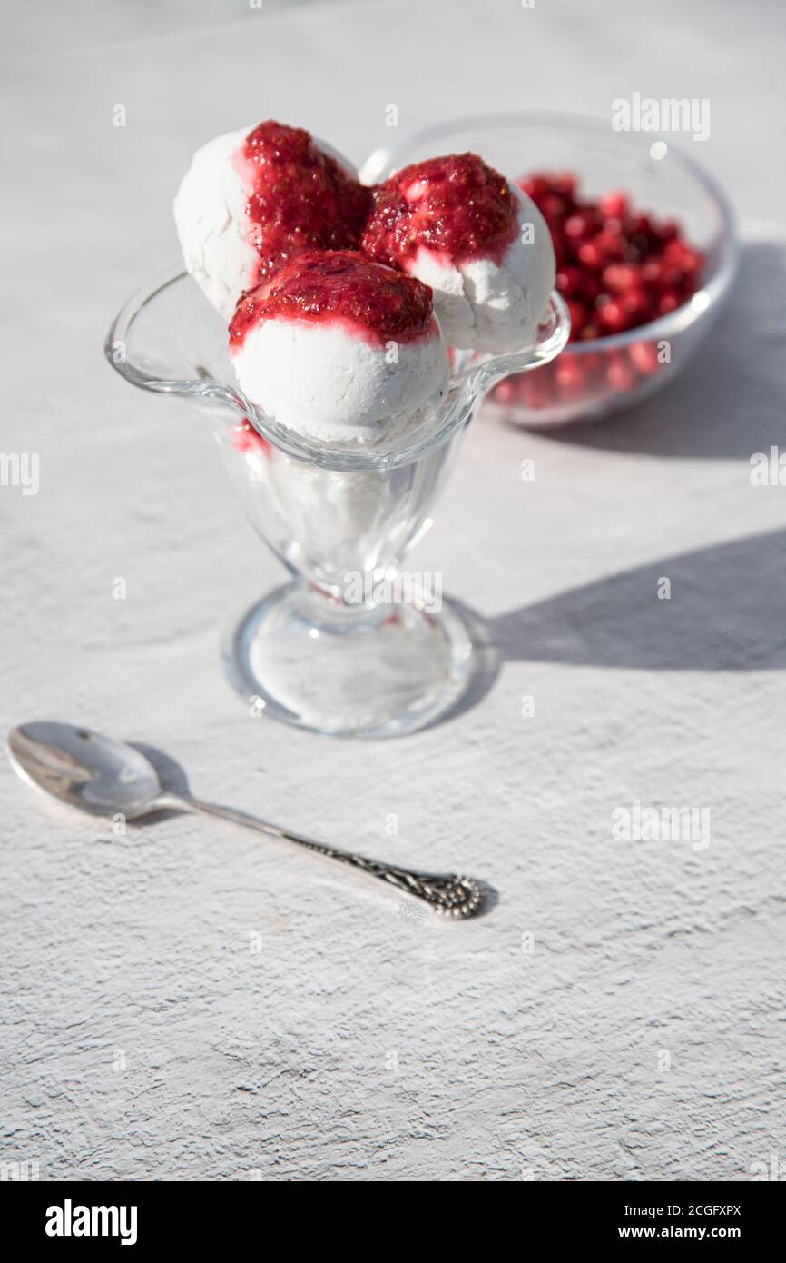 Glasschale mit drei Kugeln Eis und Beerenmarmelade auf hellem Hintergrund, Beeren in einer Glasschale, Sonnenschatten auf dem Tisch. Vertikale Ausrichtung Stockfoto
