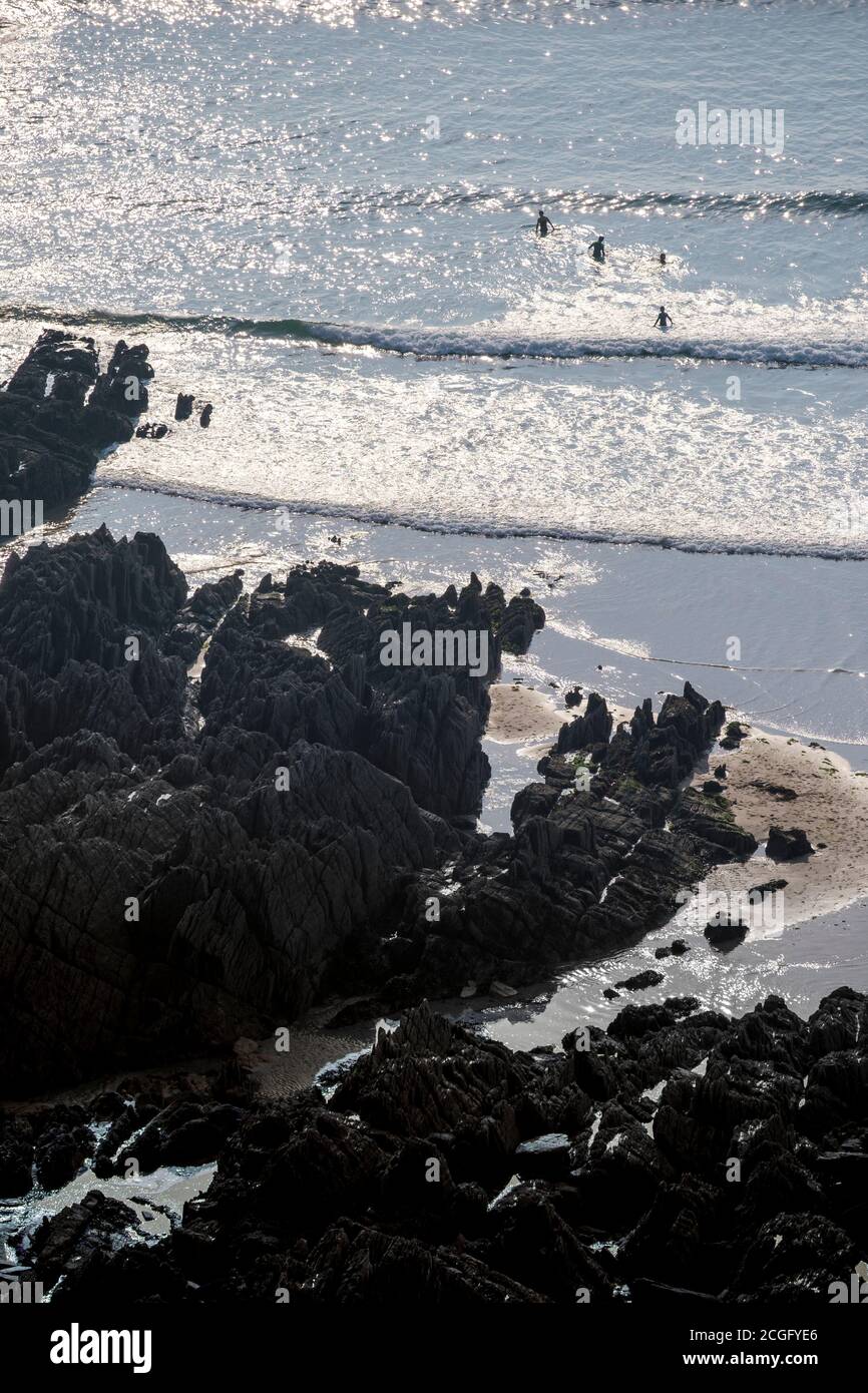 Kleine Wellen an einem Sandstrand mit großen schwarzen Felsen und die Silhouette der Menschen im Meer. Stockfoto