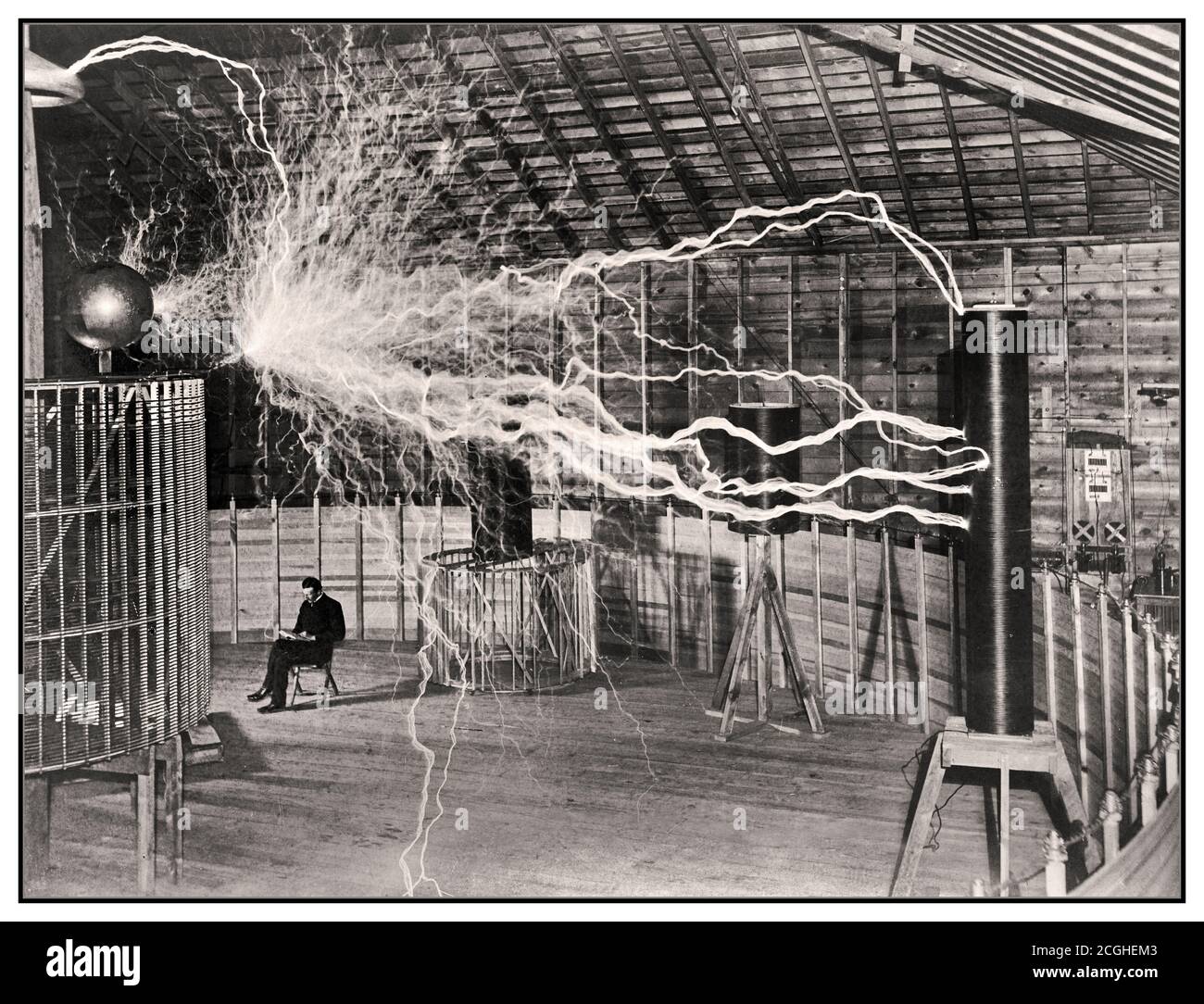 1899 Elektrizitätsexperiment Nikola Tesla serbisch-amerikanischer Erfinder in seinem Labor in Colorado Springs Dezember 1899, scheinbar sitzend neben seinem riesigen 'agnifying Transmitter' Hochspannungsgenerator, während die Maschine riesige Strombolzen produzierte. Es war ein Trick Foto von Fotograf Dickenson V. Alley; eine doppelte Belichtung. Zuerst wurden die riesigen Funken der Maschine im abgedunkelten Raum fotografiert, dann wurde die Fotoplatte bei ausgeschaltetem Gerät und Tesla im Stuhl wieder belichtet. Tesla gab zu, dass das Foto falsch ist, aber es erhielt die große Aufmerksamkeit, die er wollte. Stockfoto