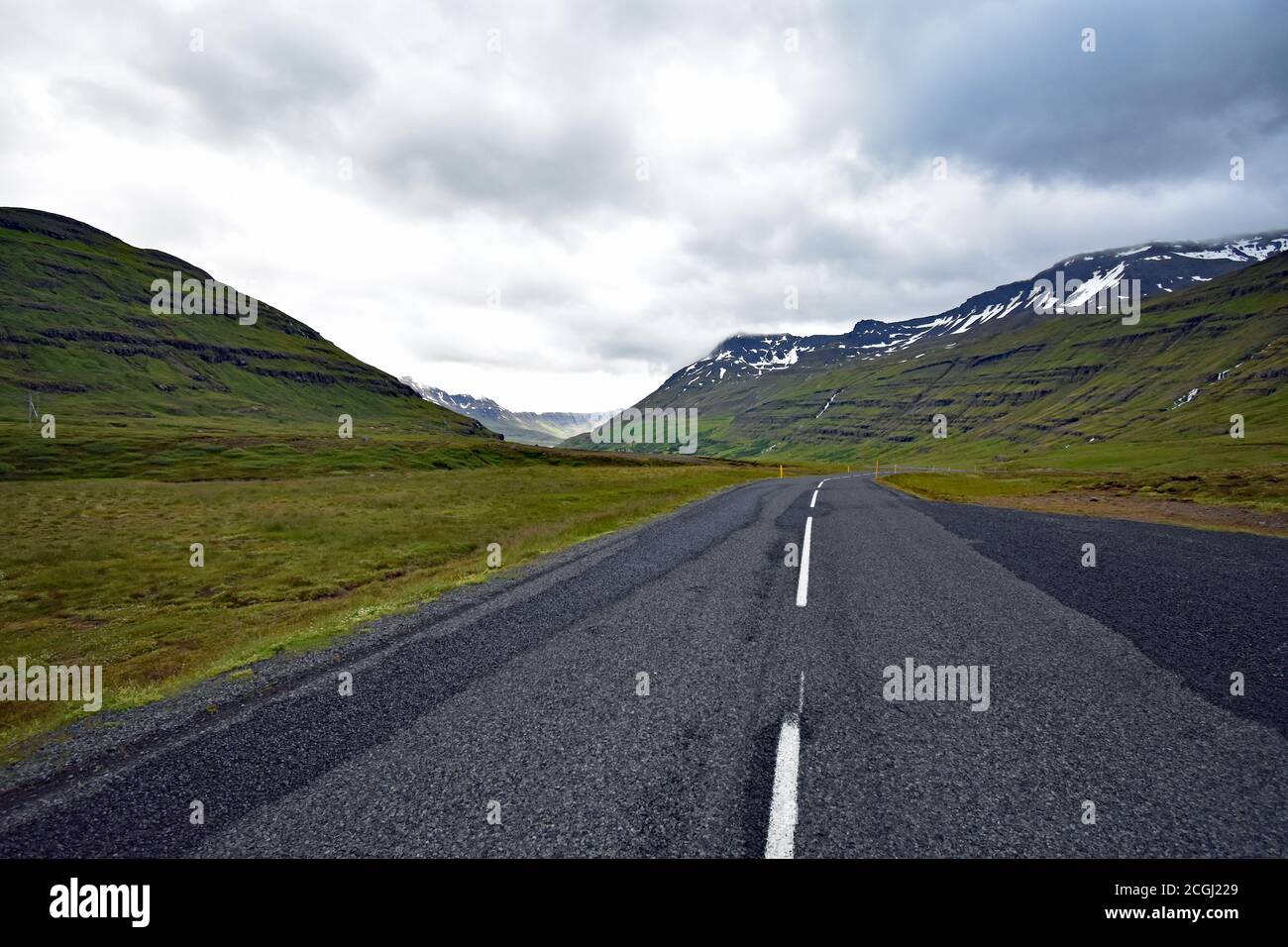 Eine Straße überquert die Berge in Seydisfjordur an einem bewölkten Tag. Die weißen Fahrbahnmarkierungen passen zum Schnee. Seydisfjardarvegur, Route 93, Island Stockfoto