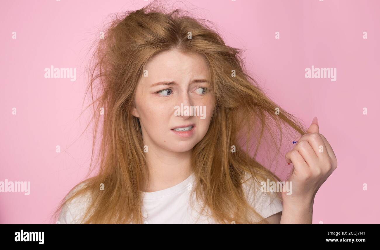 Das Konzept der trockenen leblosen Haar. Eine Frau auf einem rosa Hintergrund hält ihr zerzaust verworrenes Haar und sieht es mit einem unzufriedenen Ausdruck an. Stockfoto