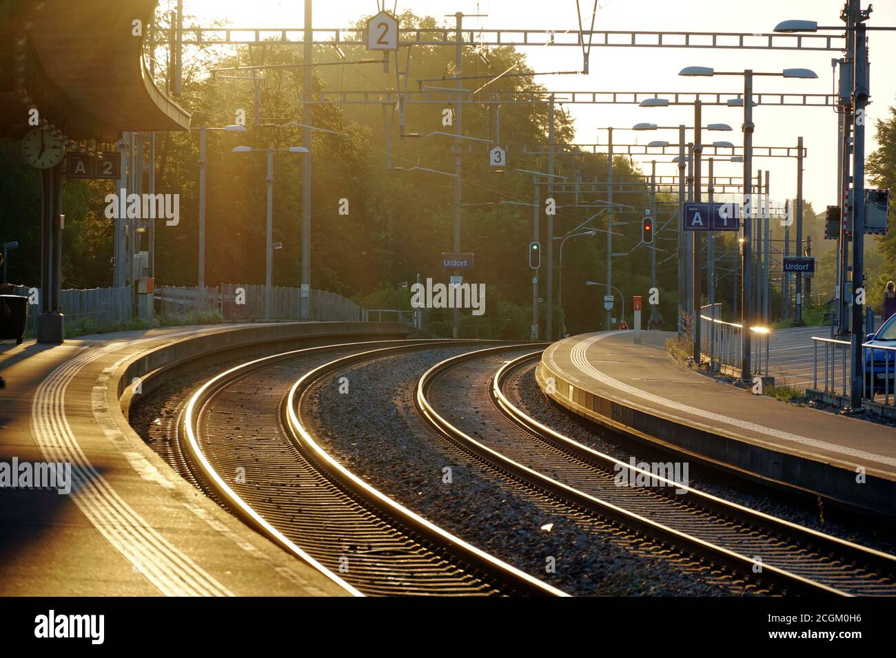 Sonnenaufgang auf dem Bahnhof Urdorf im Sommer. Das Licht fällt durch Bäume auf den Schienen und gibt ihnen goldene Reflexe. Stockfoto