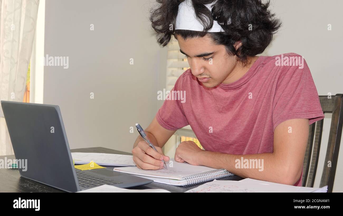 Teenager Junge der nahöstlichen Ethnie tun Remote High School in Zeiten von Covid-19, sitzen am Tisch Notizen von einem Laptop. Stockfoto