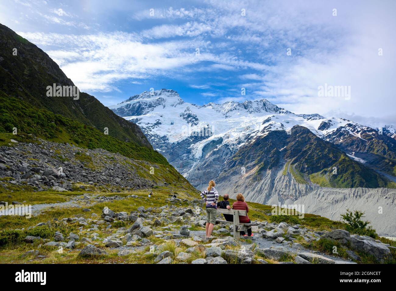 Eine dreiköpfige Familie beobachtet den mueller Gletscher am Kea Point im Mount Cook Nationalpark, die felsigen Berge und das grüne Gras Stockfoto