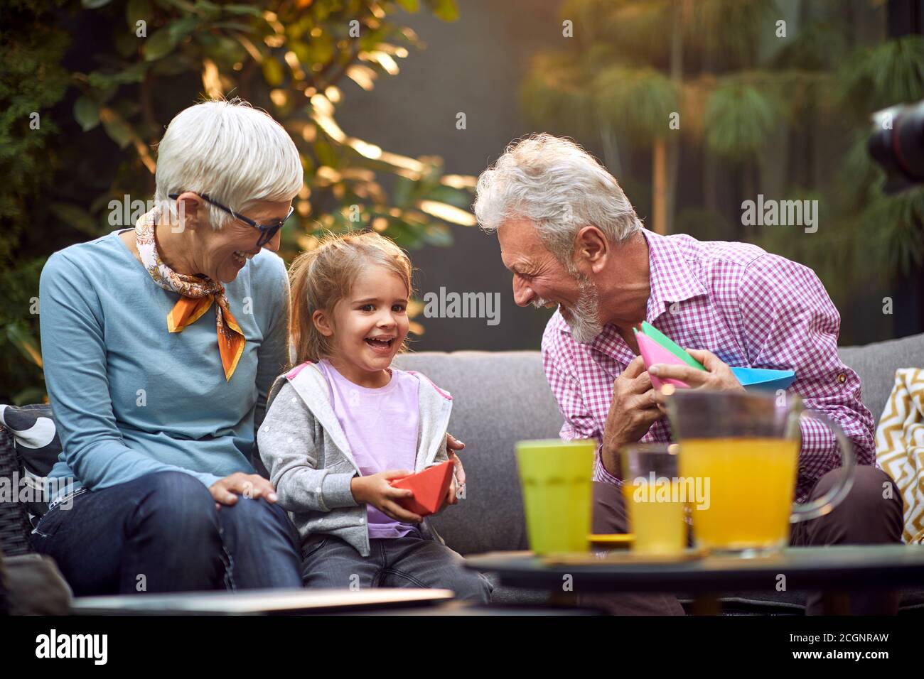 kaukasische Großeltern lachen, genießen mit ihrer Enkelin, sitzen im Freien auf dem Sofa Stockfoto