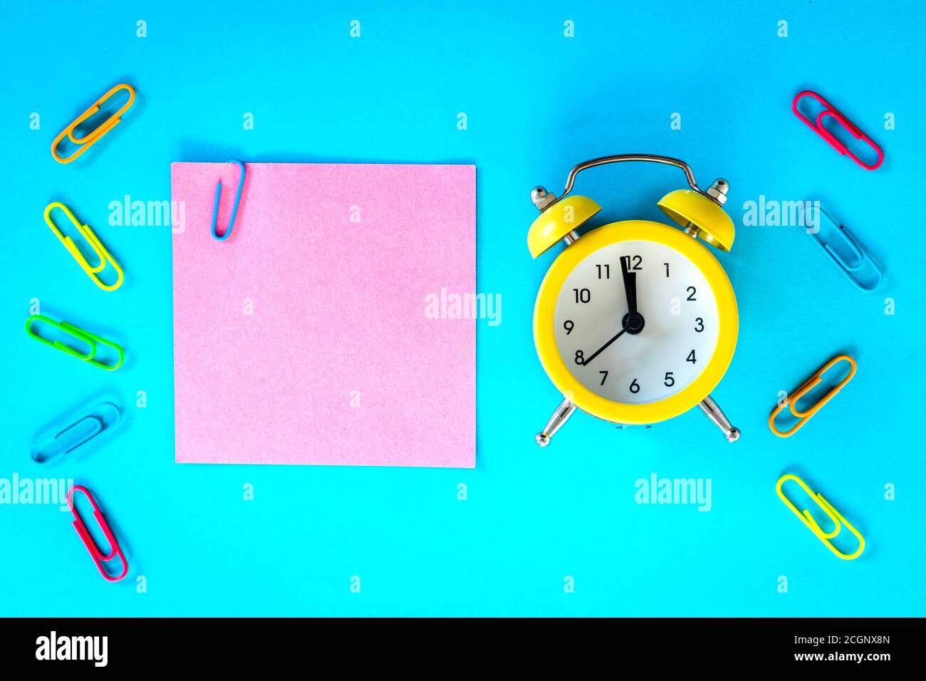 Blauer Hintergrund, pinke Karte mit Büroklammer und Platz für eine Beschriftung. Daneben steht ein Wecker für eine Stunde, fünf bis Mitternacht, zur Verfügung. Concep Stockfoto