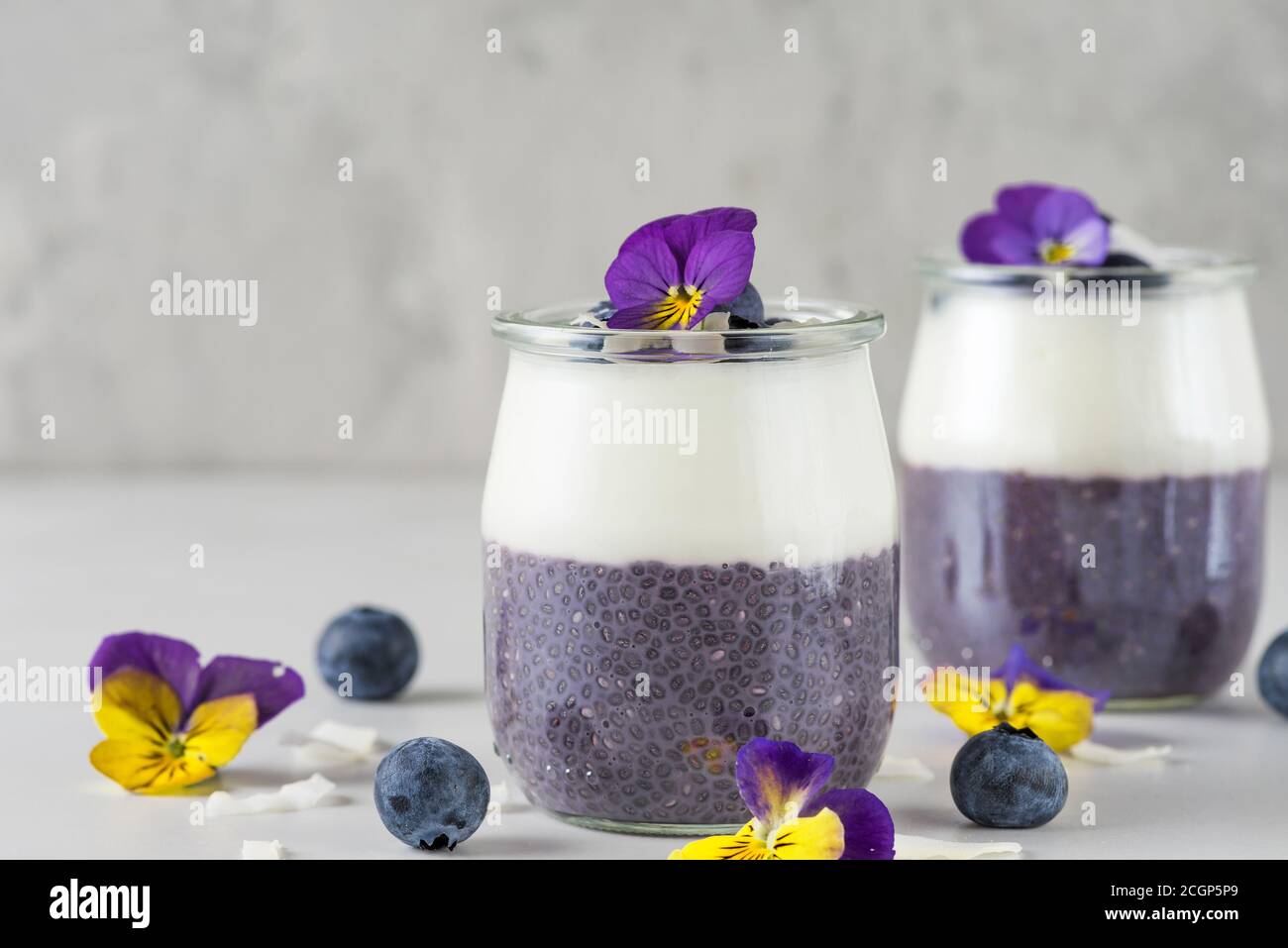 Heidelbeer Joghurt Parfait mit Chia Pudding, Kokosjoghurt, frische Beeren und Blumen in Gläsern für gesunde Ernährung Frühstück auf weißem Marmortisch mit Stockfoto