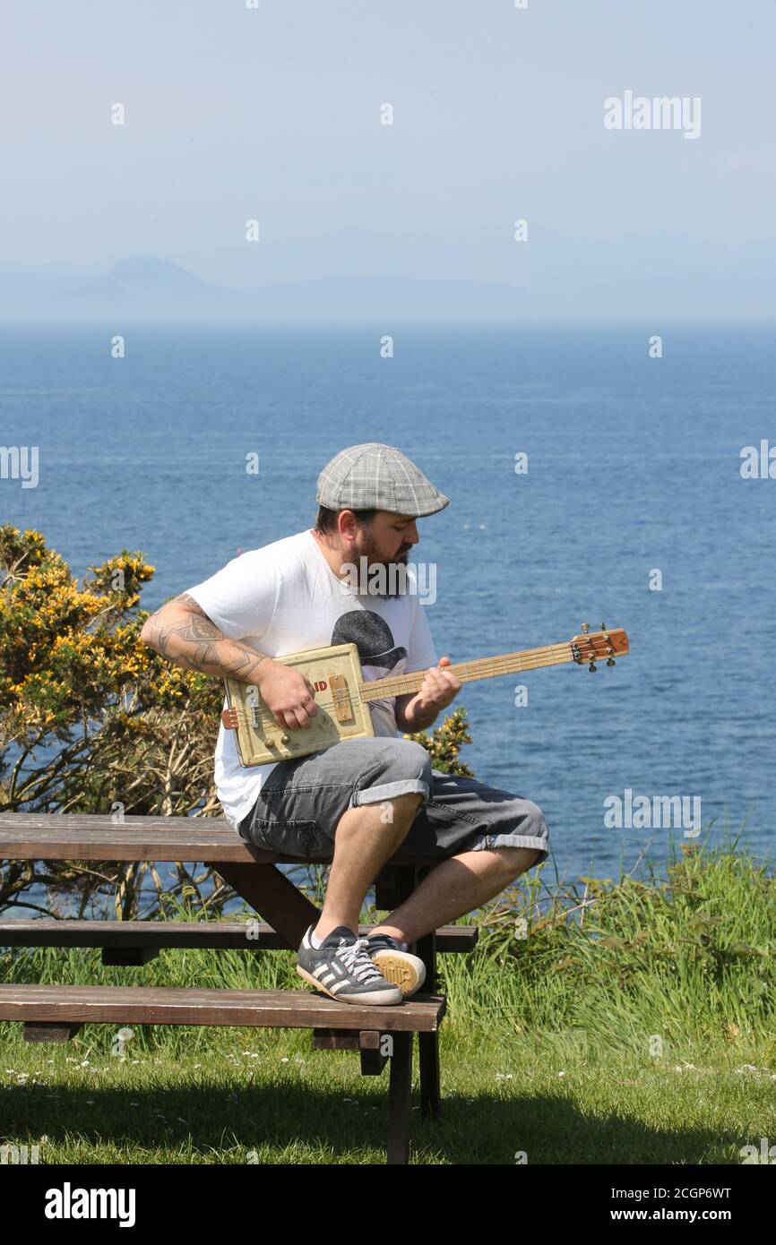 Dunure, Ayrshire, Scotland, UK 16 May 2019 : Nick Wild, der in einem umgebauten Van durch Großbritannien reist und seinen Lebensunterhalt mit handgemachten Gitarren bestreiten kann. Müde von seinem Job als Stahlarbeiter in Sheffield & mit einem Interesse an Musik & Holzbearbeitung verkaufte er sein Haus, kaufte einen alten Lieferwagen, verwandelte es in Wohnunterkunft & mobile Werkstatt. Saß auf einer Bank und spielte seine handgemachte Gitarre aus einer alten erste-Hilfe-Box mit dem Ayrshire Coast & Dunure Castle im Hintergrund Stockfoto