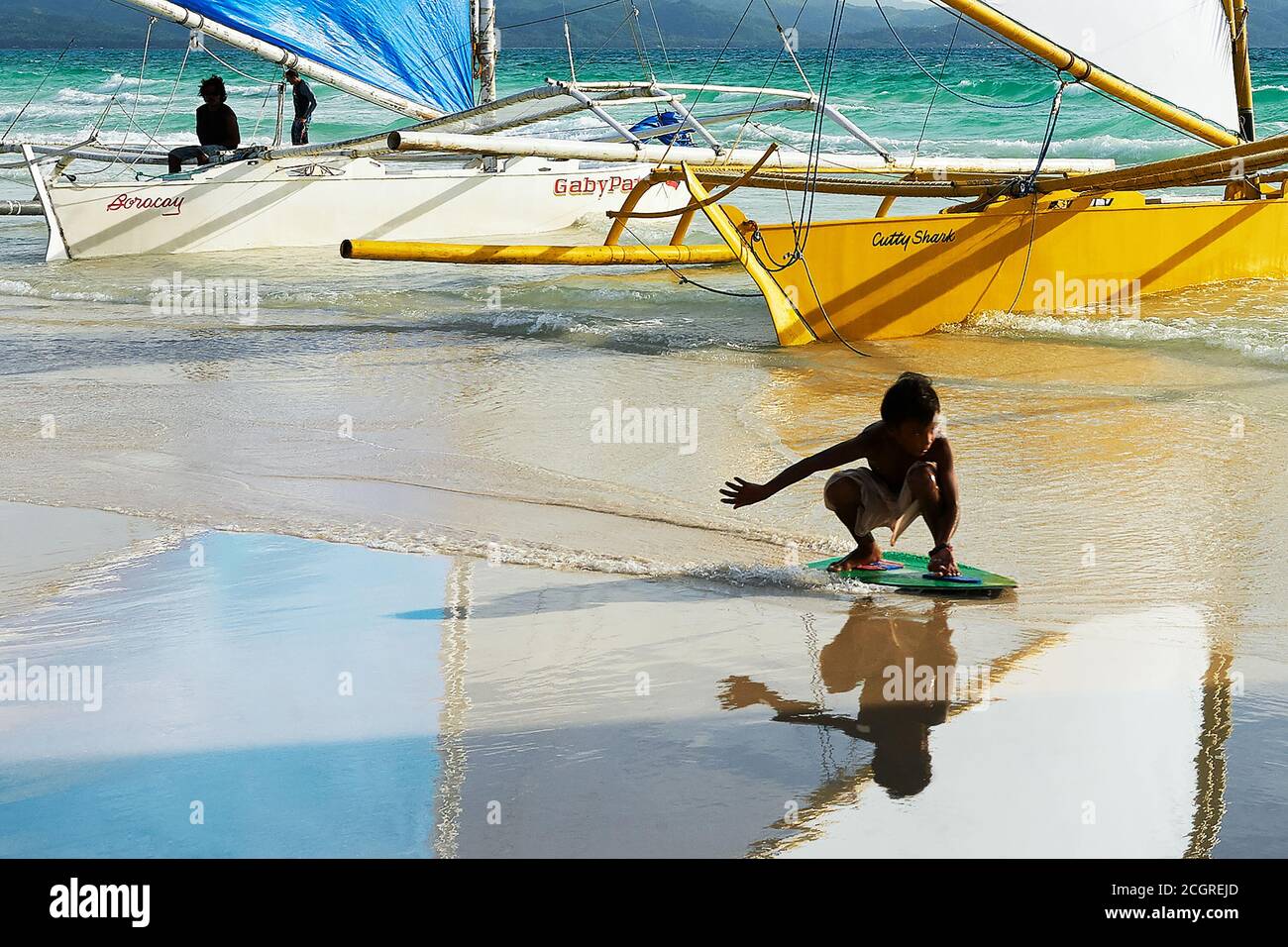 Bunte Segelboote und ihre Spiegelungen am White Beach auf Boracay Island, und die Silhouette eines Jungen, der auf einem Skim-Board auf den Philippinen reitet Stockfoto